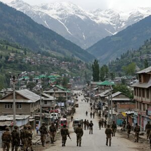 A large contingent of armed Indian soldiers marches down a street in a Kashmiri town, with snow-capped mountains visible in the background.
